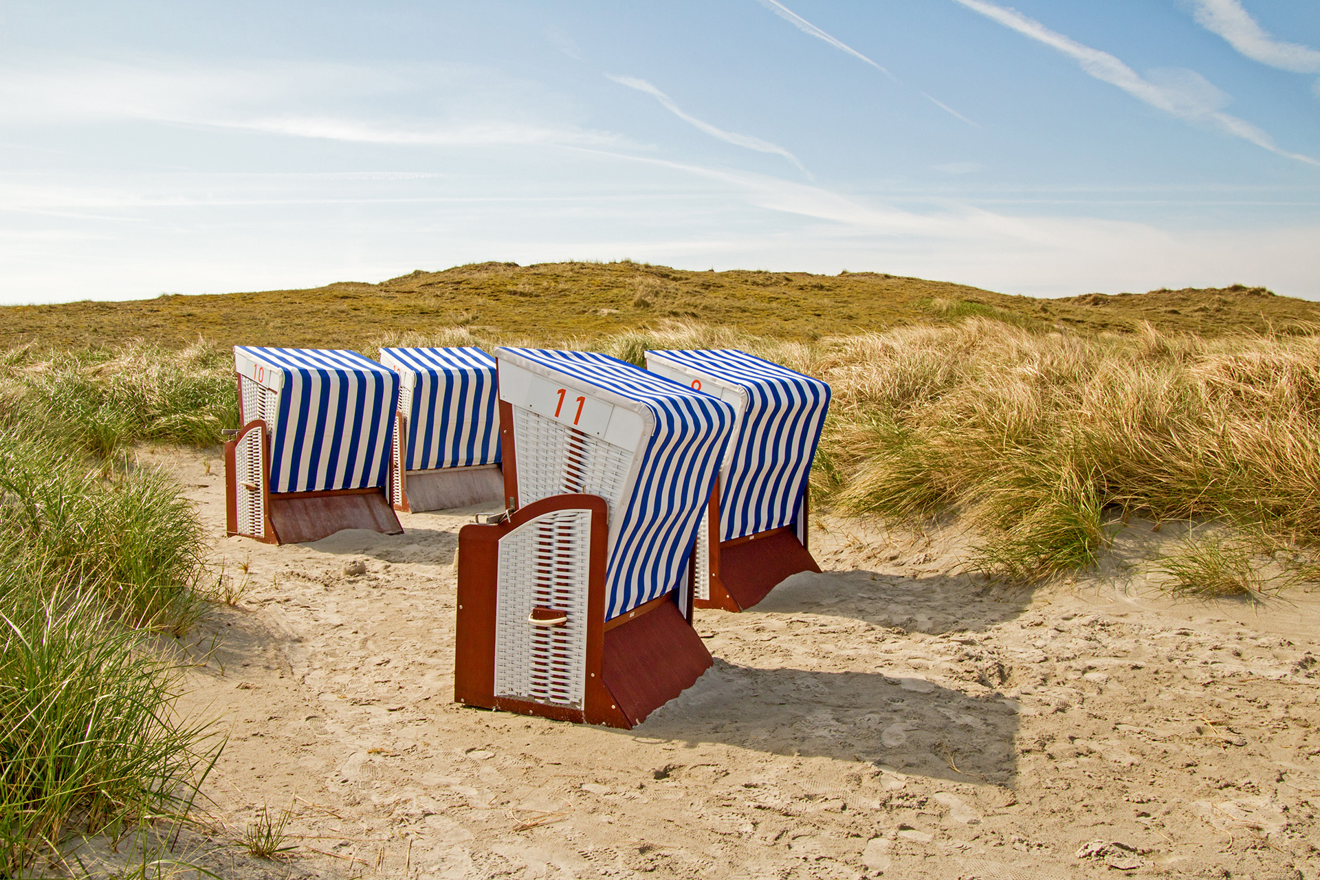 Vier blau-weiß gestreifte Strandkörbe stehen auf Sandboden in Mitten der Dünen, der Himmel ist blau.