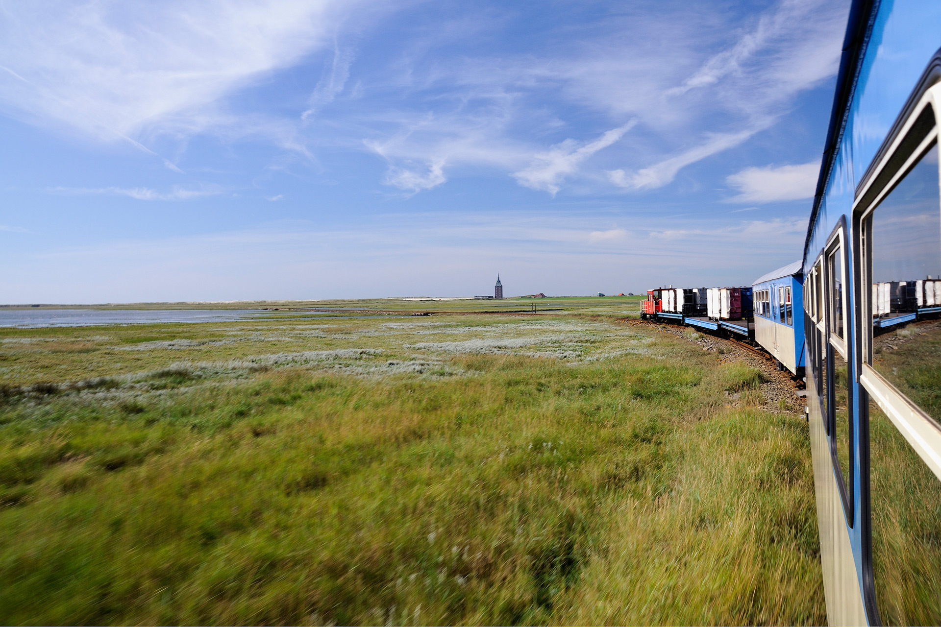 Ein Blick aus dem Zugfenster: Man sieht grüne Wiesen, viel Himmel und einen Teil des Zuges selbst. Am Horizont in Bildmitte ragt der Westturm von Wangerooge.